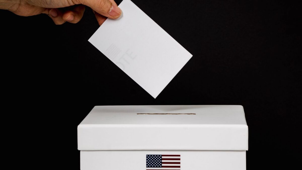 Hand placing a ballot into a ballot box marked with the word Vote next to a United States flag.