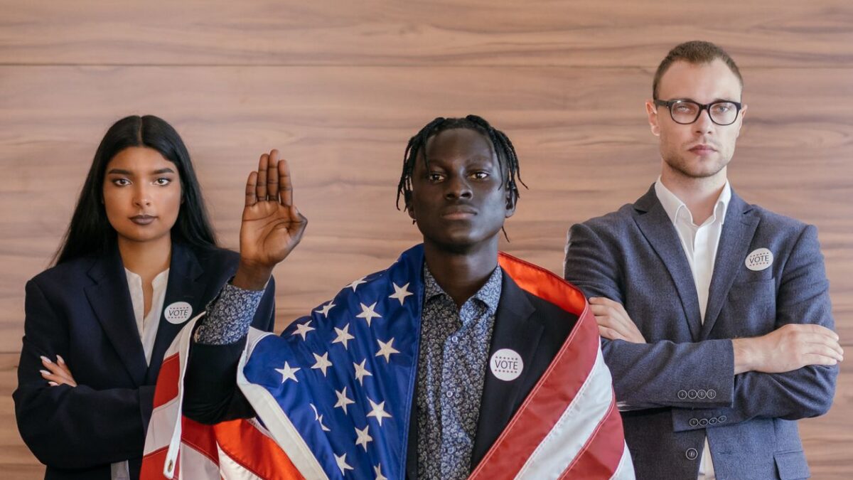 Three diverse people at a podium, one with a raised hand taking an oath, draped in the United States flag